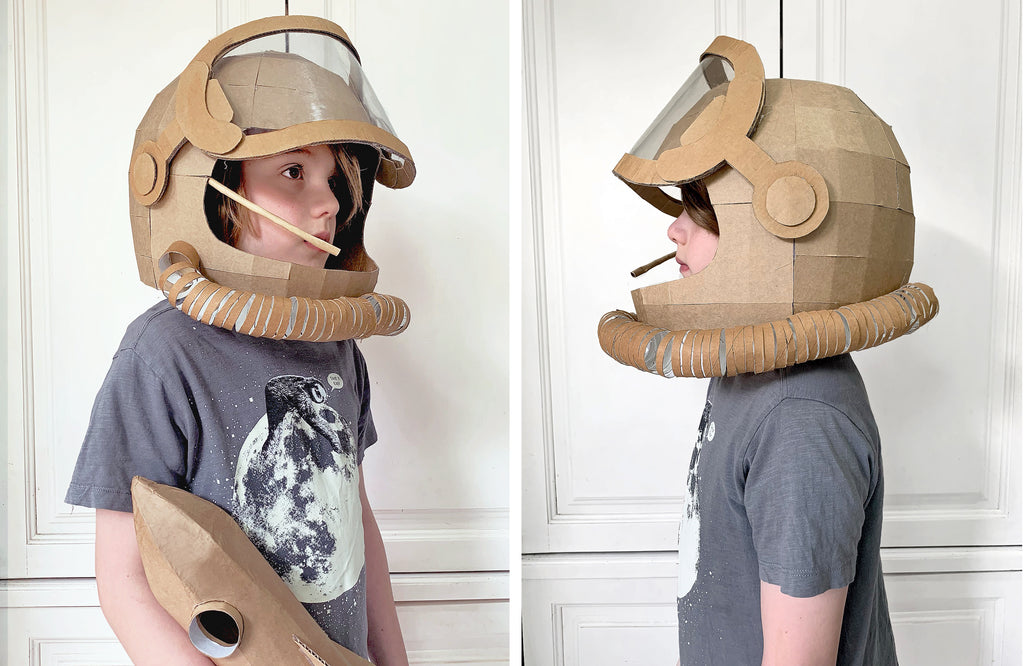Child wearing a cardboard astronaut helmet with visor and space suit design, standing against a white paneled wall.