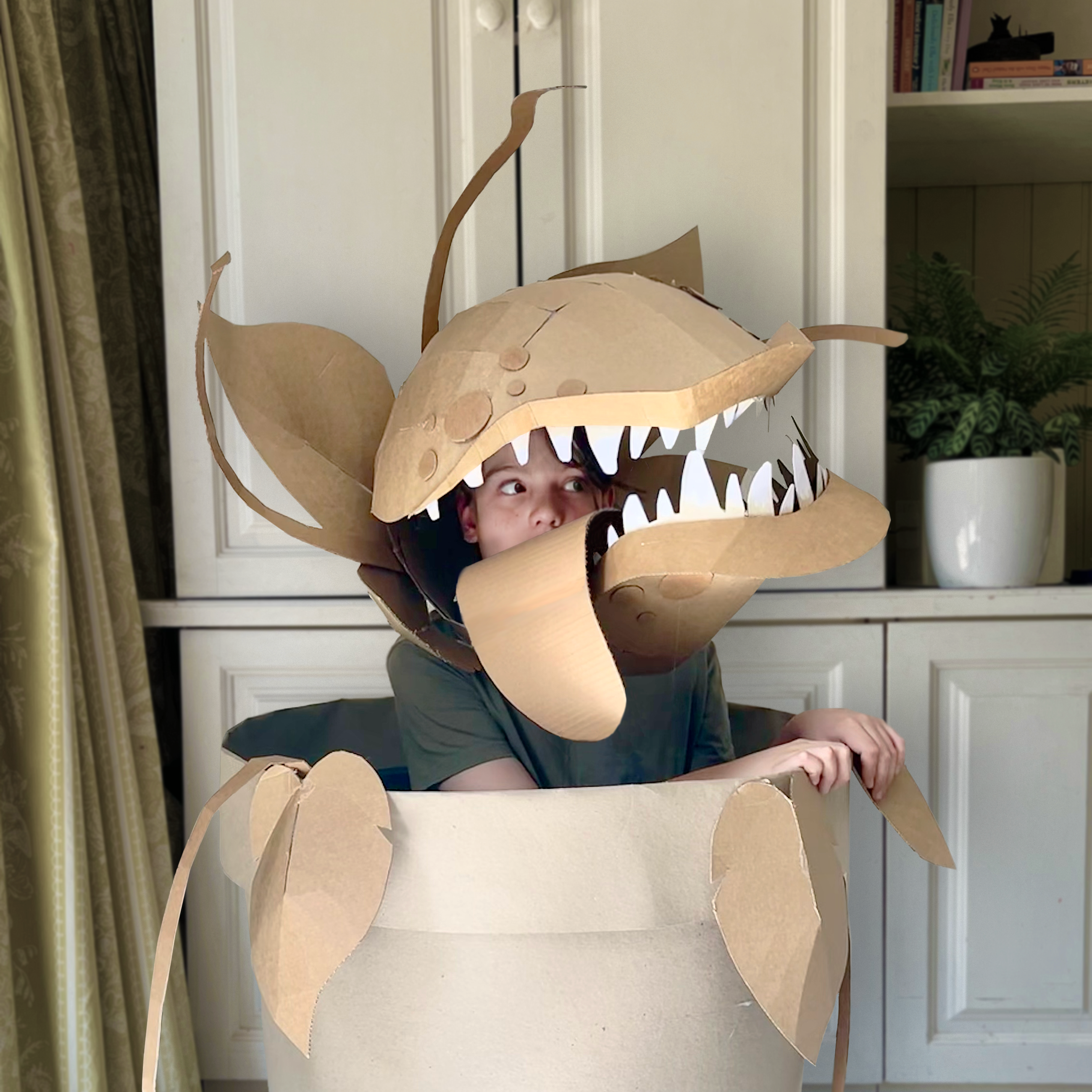 Person wearing a cardboard man eating plant costume in the style of Audrey 2 inside a room with white cabinets.