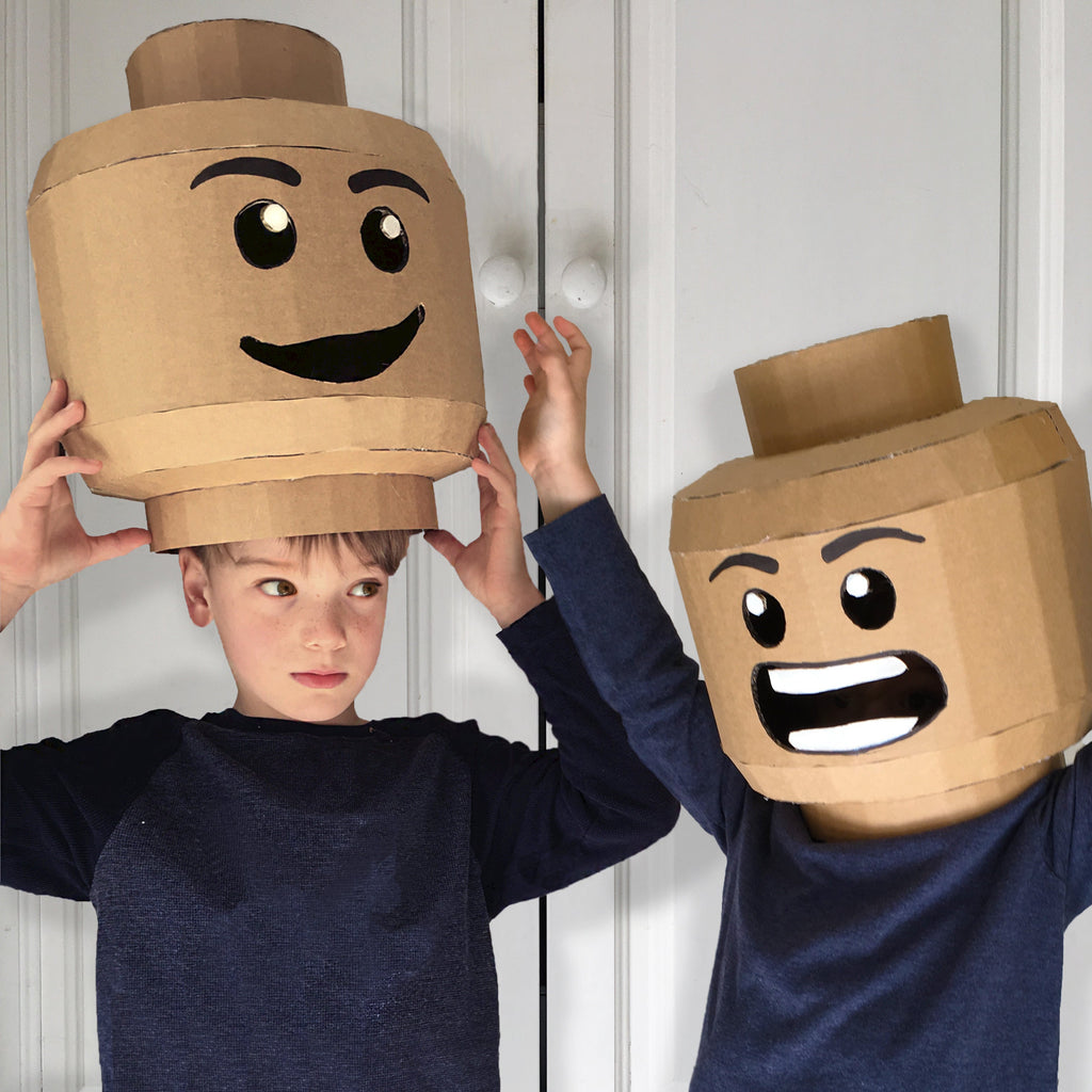 Two children wearing cardboard costumes inspired by LEGO heads against a white panelled wall.