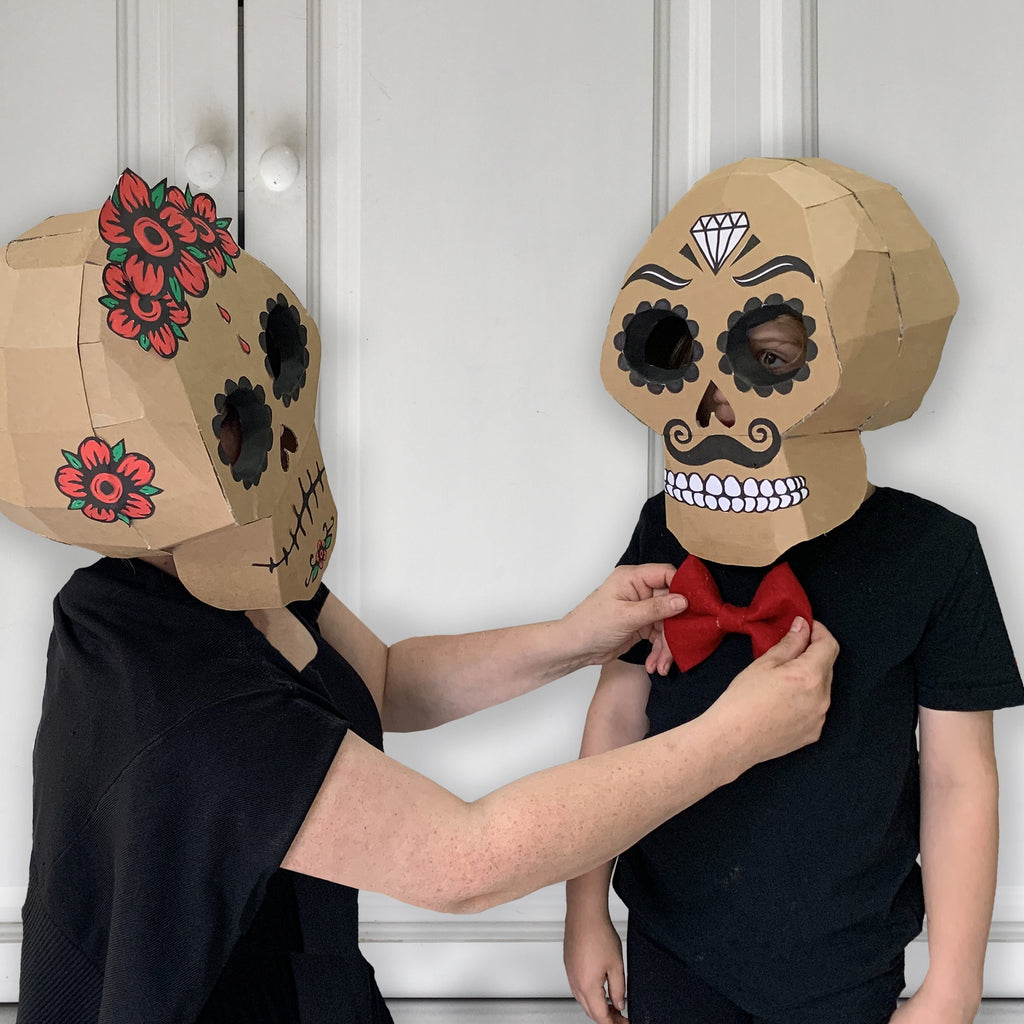 Two people wearing cardboard sugar skull mask costumes with decorative elements against a white wall.