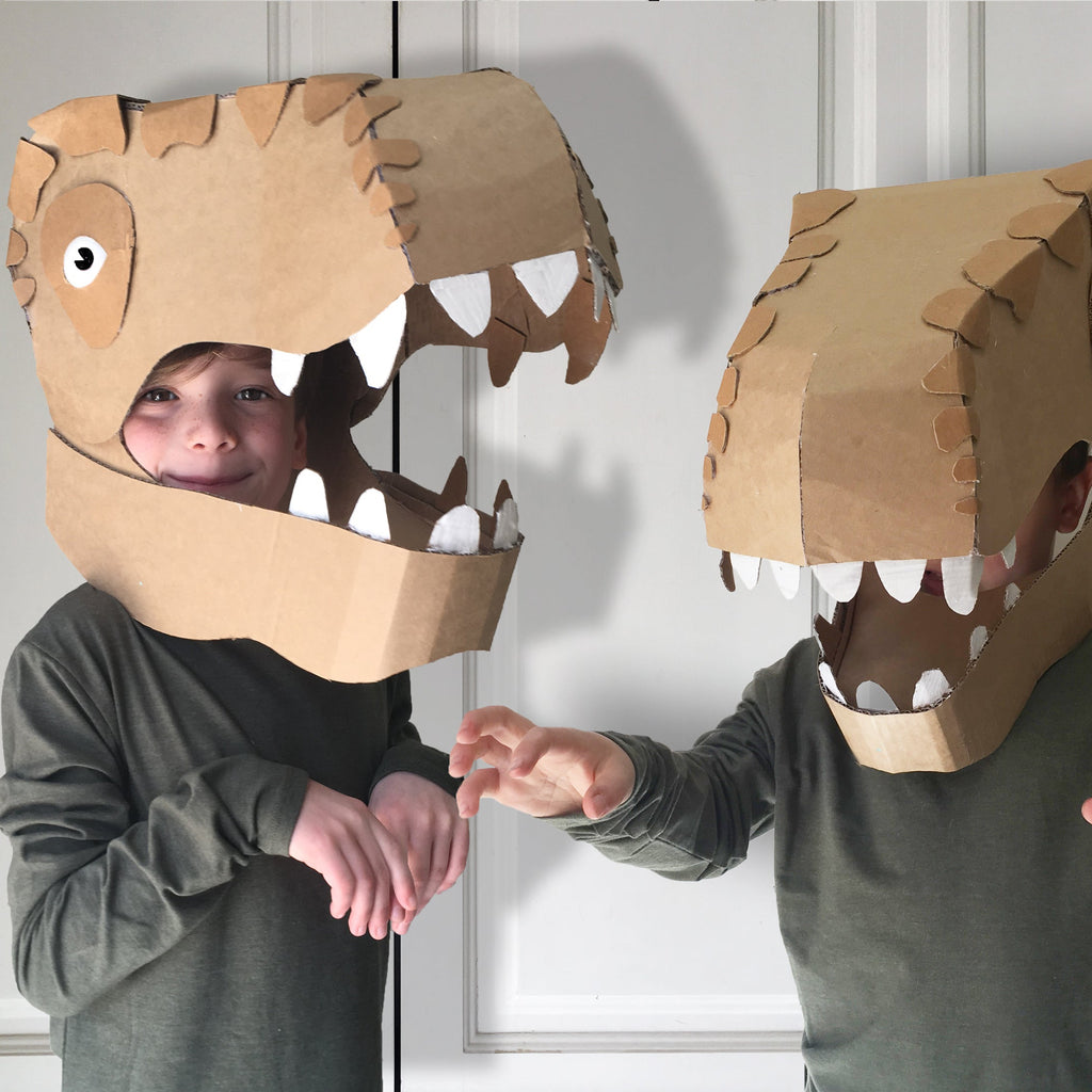 Two children wearing cardboard dinosaur masks against a white wall.