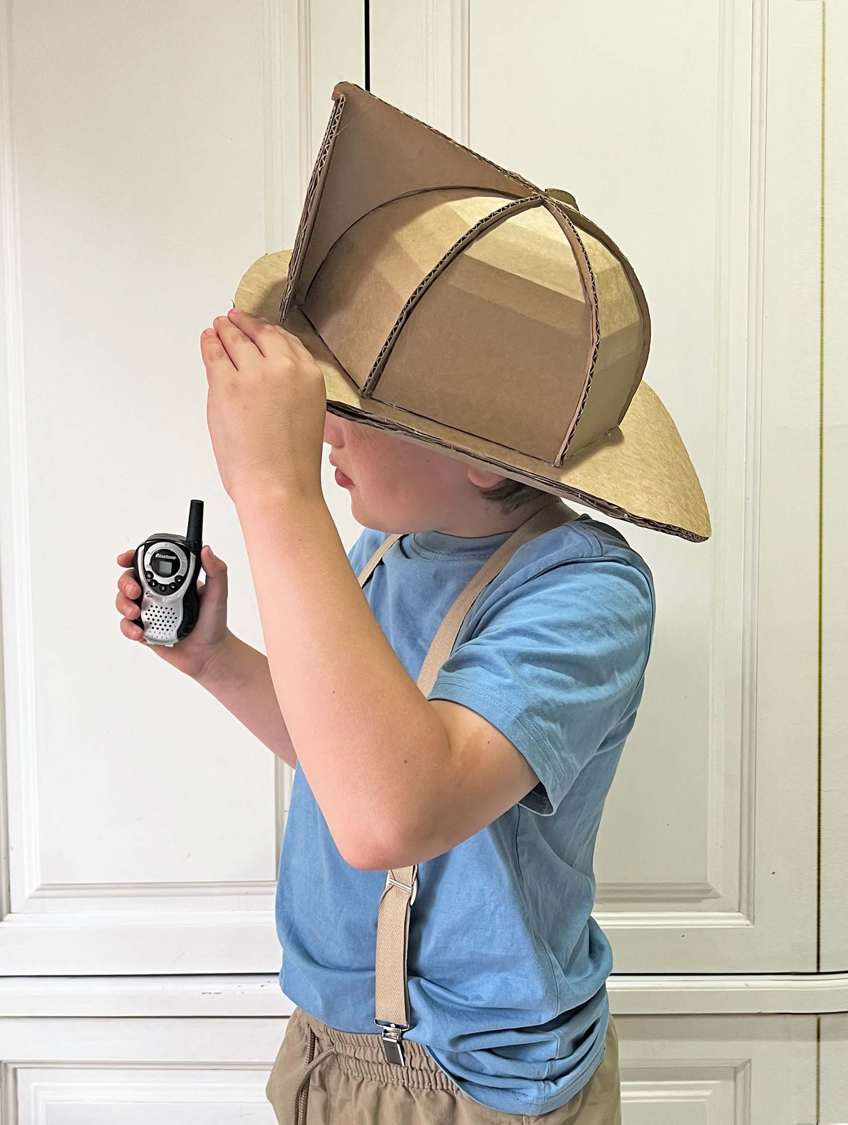 Child wearing a cardboard fireman helmet hat and holding a walkie-talkie indoors.