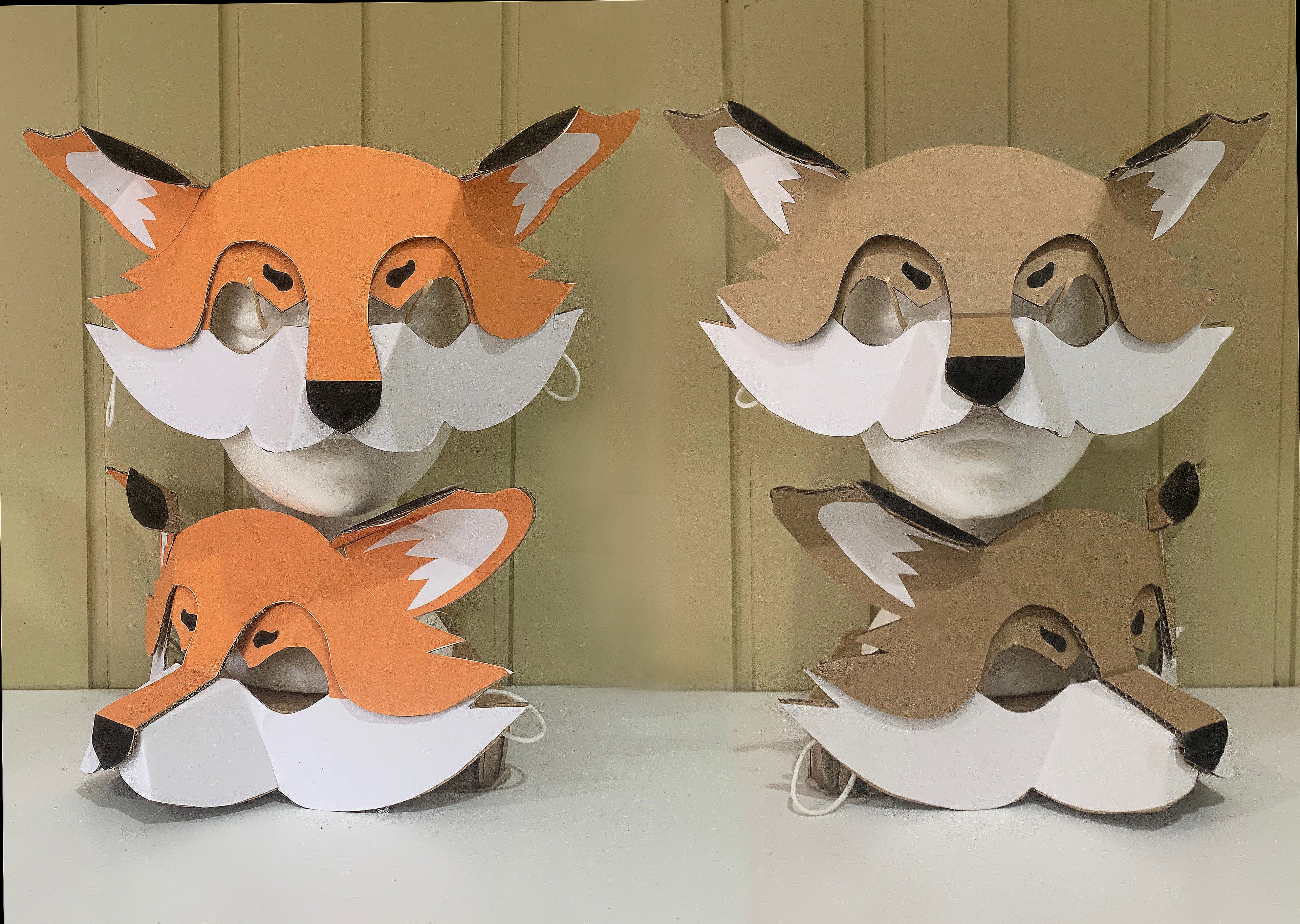 Two cardboard animal masks, one with an orange fox face  and the other with natural cardboard tones. Both using paper overlays over cardboard to create a clever fox mask.

